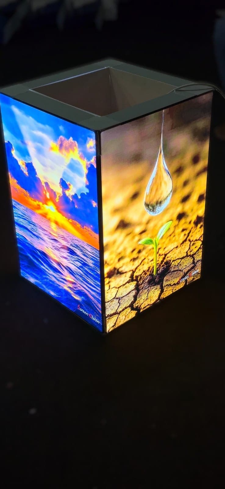 Backlit lightbox cube with printed sunset and water-drop graphics, lit at night